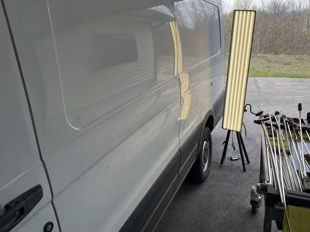 A dent repair light board on a stand beside a white cargo van in a parking lot, its stripes showing small hail dents on the side panel, a cart of repair rods next to it