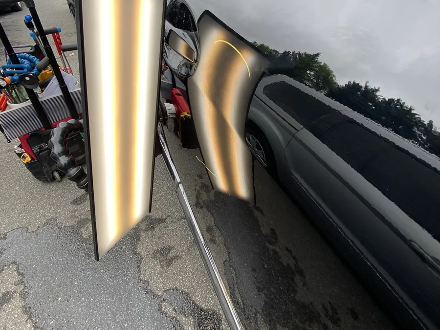 A dent light board set up against a vehicle's side panel in a parking lot, the stripes bending around a dent